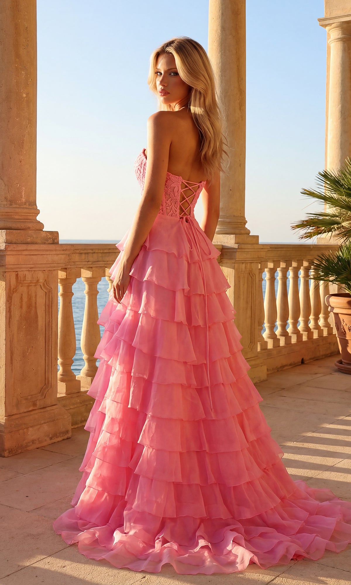 A woman wearing the Strapless Long Ruffled Prom Gown: PromGirl 250001 with a sheer corset-style bodice stands on a stone balcony at sunset, overlooking the sea with columns and potted palm in the background.